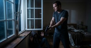 A man points a handgun at a hooded intruder surrendering on a bedroom floor next to a broken window