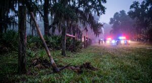 Pickaxe leaning against a fence in a grassy field with flashing police vehicles in the foggy background